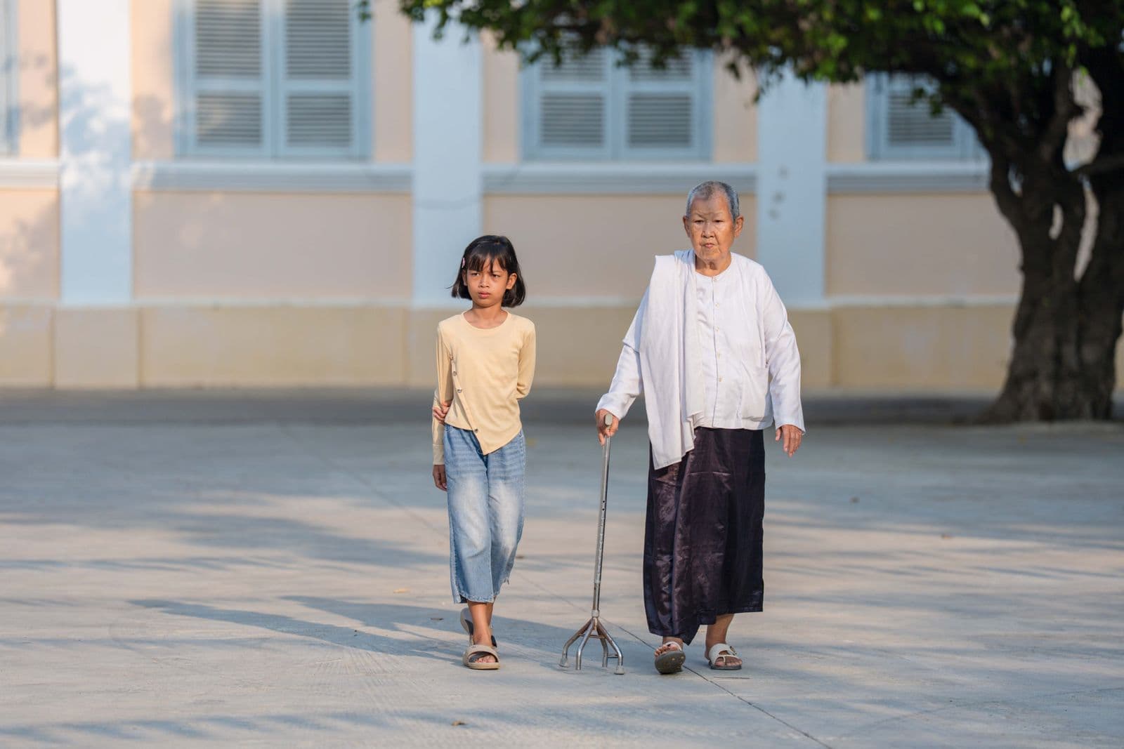An elderly Thai woman walking outdoors hand in hand with her young granddaughter