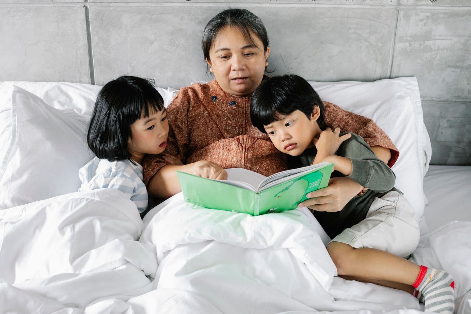 An older Thai woman resting in bed reading with her grandchildren at home
